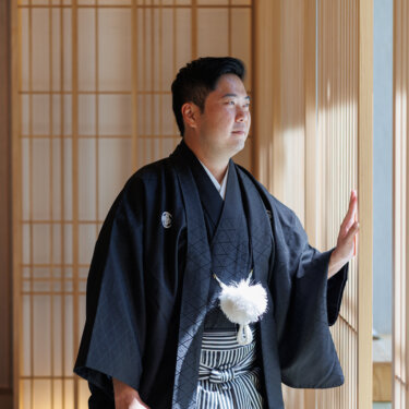 Groom in black montsuki hakama holding a folding fan gazes out past the lattice screens, a solo portrait from a Japanese wedding photoshoot