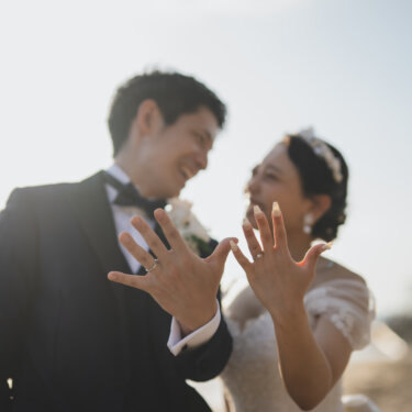 Smiling couple showing off their wedding rings with spread fingers by the sea, a fun ring pose for pre-wedding photos near Noma Lighthouse