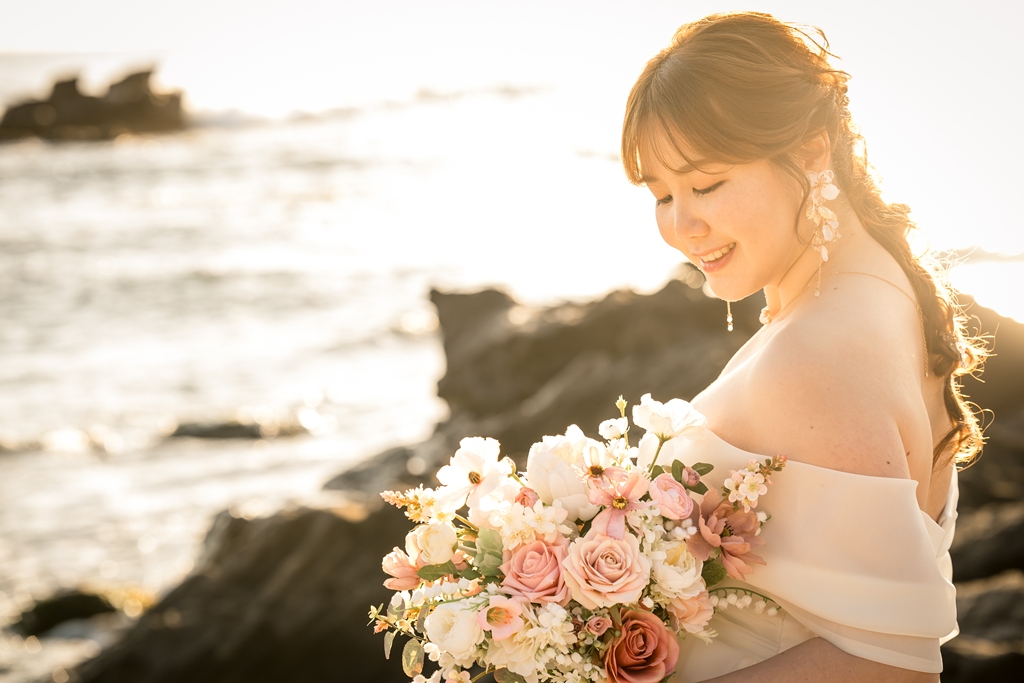 Close-up of a smiling bride in an off-shoulder wedding dress holding a bouquet in the warm sunset light on Jogashima