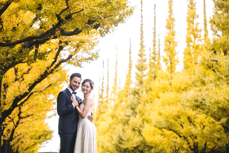Bride in a wedding dress and groom in a tuxedo standing close together under golden ginkgo trees in Tokyo