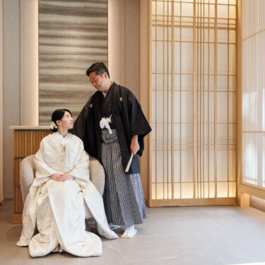 A bride in shiromuku sits in an armchair while the groom in montsuki hakama stands beside her in a softly lit studio lounge, a classic kimono wedding photo