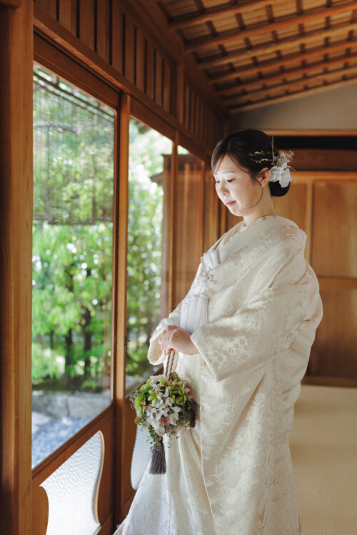 Bride in the NS102 shiromuku with fresh flower hair accessories lowering her eyes by a wooden window with a bouquet, a pre-wedding photoshoot in Japan