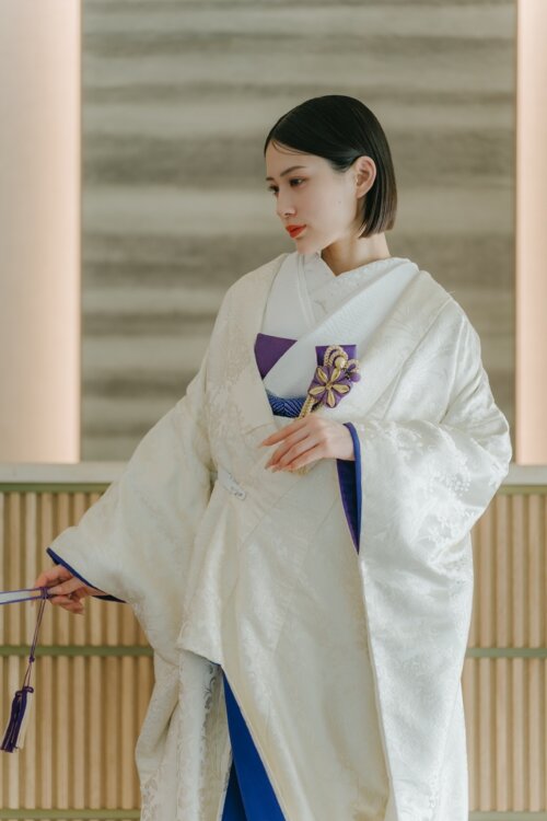 Bride in a shiromuku looking down with a purple hakoseko and fan beside a wooden counter in our studio, the blue under-kimono standing out in this kimono wedding photo in Japan (AR102).