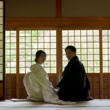 Bride in shiromuku and groom in montsuki sitting on tatami in front of glowing shoji screens and looking back over their shoulders, a beautiful back-view pose for a pre-wedding photoshoot