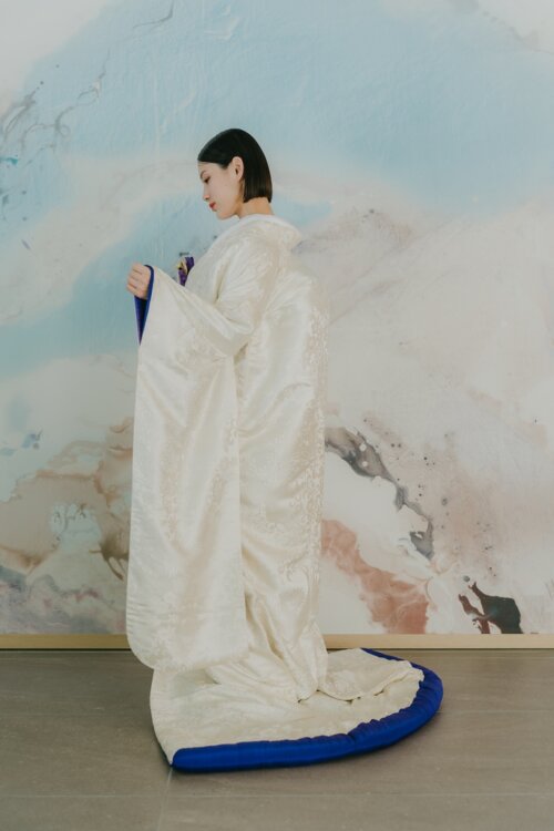 Side view of a bride in a shiromuku spreading the blue-trimmed hem in front of a watercolor-style wall, Japan wedding photography with a striking accent color (Shiromuku AR102).
