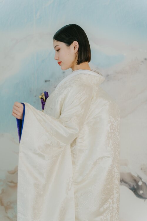Profile of a bride in a shiromuku glancing down to reveal a vivid blue under-kimono before a pale blue art wall, a pre-wedding photoshoot in Japan in our studio (AR102).