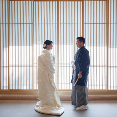Bride in a long-trained shiromuku and groom in montsuki hakama face each other across a bright room lined with lattice screens, Japan wedding photography