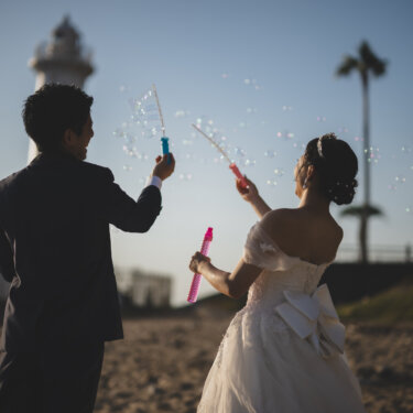 Bride and groom blowing soap bubbles on the beach with the lighthouse and palm trees behind them, a playful prop idea for Chita Peninsula wedding photography