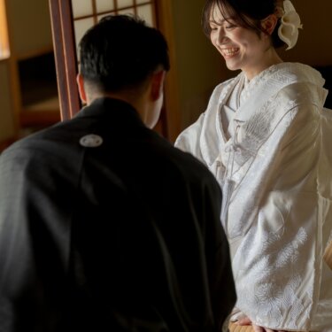 Smiling bride in shiromuku with an orchid hair ornament kneeling on tatami facing the groom by a shoji window, a warm indoor moment from their kimono wedding photos