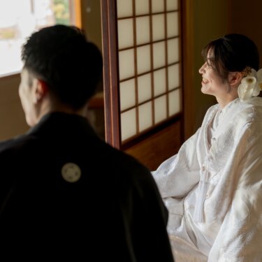 Bride in shiromuku sitting by the shoji screen and smiling at the groom in black montsuki seen from behind, soft window light in a tatami room for Japan wedding photography