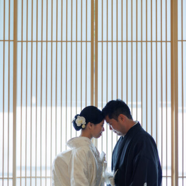 Backlit by tall lattice screens, the bride in shiromuku and groom in montsuki hakama lean their foreheads together, a quiet kimono wedding photo pose