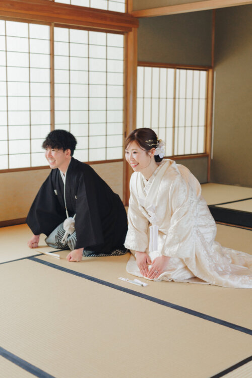 Groom in montsuki hakama and bride in shiromuku kneeling side by side and laughing in a tatami room lit through shoji screens, natural Japan wedding photography