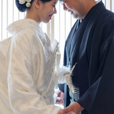 Bride in shiromuku with a white orchid hair ornament and groom in black montsuki hakama touch foreheads by lattice screens in a Japanese studio wedding photoshoot