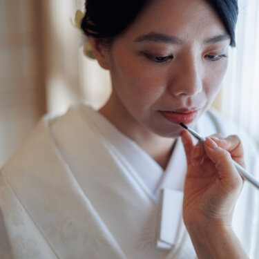 A bride in a white shiromuku kimono has her lipstick applied with a brush by the window, a getting-ready moment before her kimono wedding photoshoot