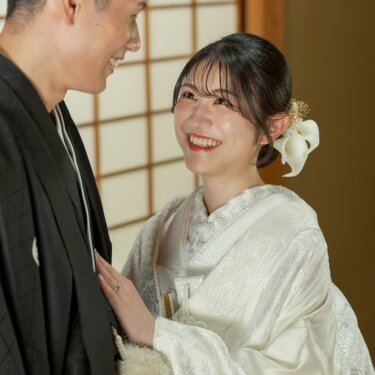 Bride in shiromuku with a white flower hair ornament laughing up at the groom with her hand on his chest, a close-up couple portrait in a tatami room for their wedding photos