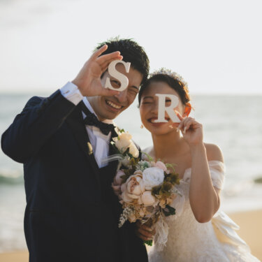 Laughing couple holding up wooden initial letters to their faces by the waves, with a peony bouquet in a seaside pre-wedding photoshoot