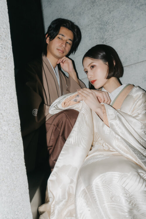 Bride in a shiromuku with a shimmering crane pattern seated beside the groom in a brown montsuki and hakama against a concrete wall, modern kimono wedding photos in Japan (DR101).