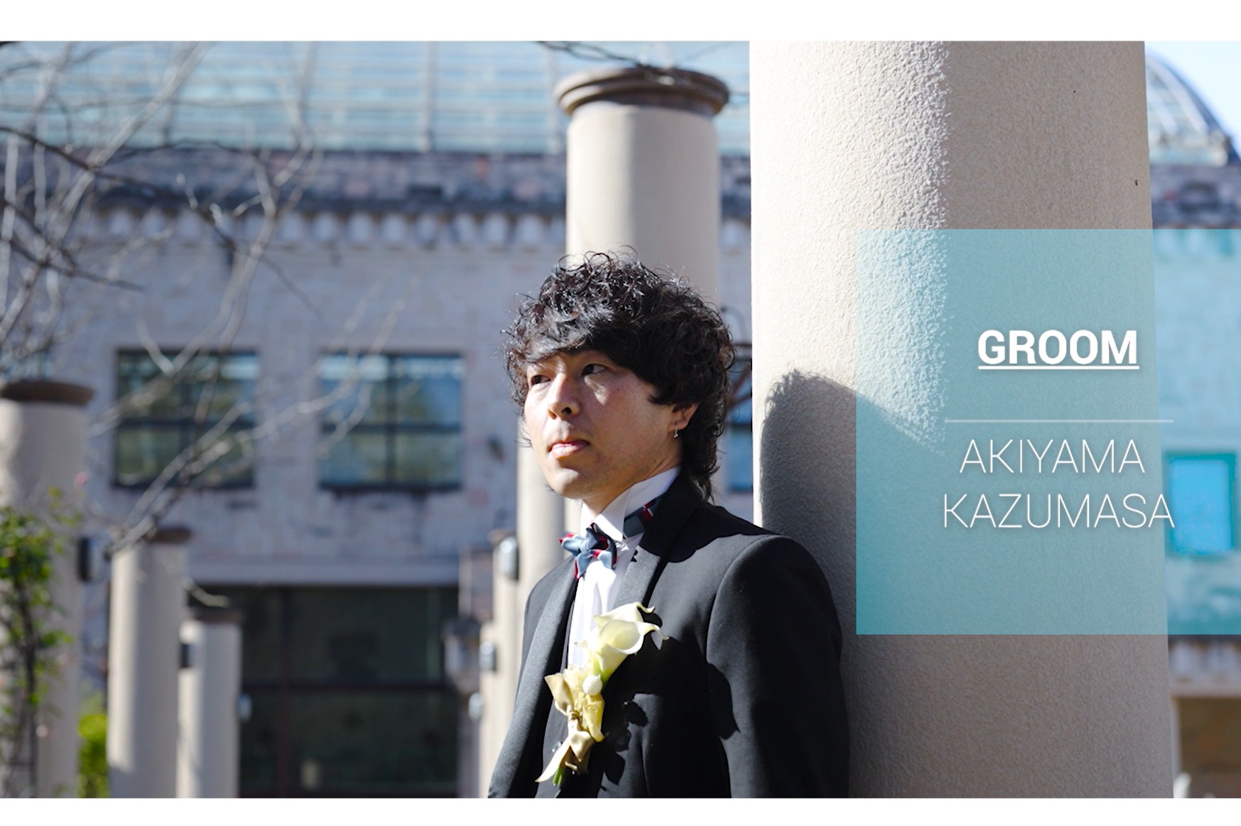 Video still reading 'GROOM AKIYAMA KAZUMASA', groom wearing a white boutonniere beside historic pillars.