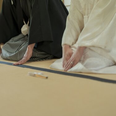Close-up of the bride and groom kneeling on tatami with their hands placed together in a traditional bow, folding fans set before them, a respectful detail from a kimono wedding photoshoot