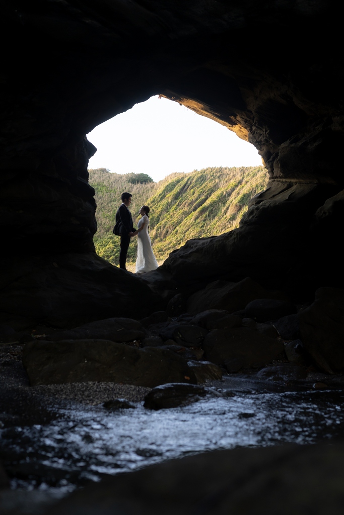View from inside a sea cave on Jogashima looking out to a couple standing on the bright shore, dramatic wedding photography near Tokyo