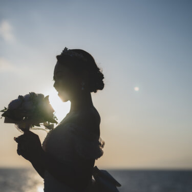 Silhouette of the bride holding her bouquet up against the setting sun over the sea, a backlit bridal portrait for Japan wedding photography