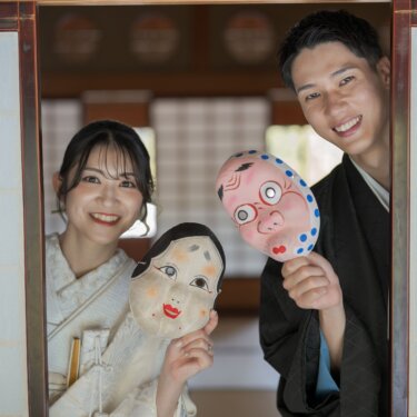 Bride and groom peeking through an open shoji window holding Okame and Hyottoko masks beside their faces, a playful props idea for pre-wedding photos in Japan