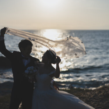 Groom lifting the bride's veil in the wind as they stand on the shore at sunset, a romantic veil pose for beach wedding photos in Aichi