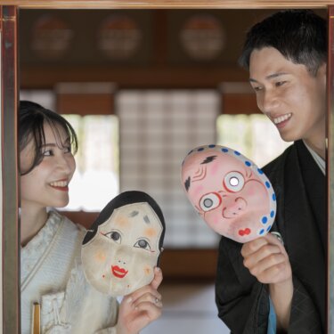 Couple in kimono laughing together while holding Okame and Hyottoko masks at a shoji window in Daisen Park Japanese Garden, a fun prop shot from their wedding photoshoot