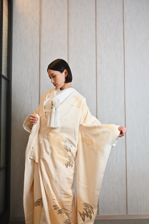 Bride in a shiromuku opening one sleeve and looking down before a wooden wall, a studio pose that shows off the gold feather pattern, kimono wedding photos in Japan (Shiromuku AB101).
