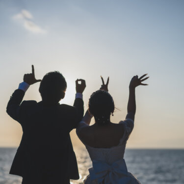 Silhouettes of the bride and groom making L-O-V-E hand letters against the sunset sky, a creative pose idea for seaside wedding photography