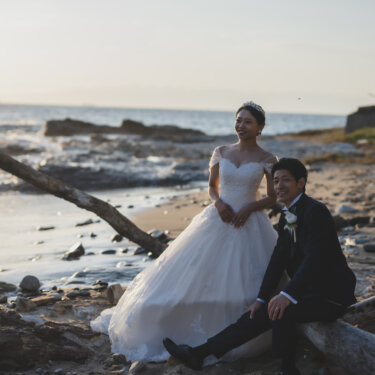 Bride standing and groom sitting on driftwood on the pebbly shore in the evening light, relaxed beach wedding photos on the Chita Peninsula