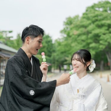 Groom in montsuki feeding the bride in shiromuku a stick of tricolor dango on a tree-lined path in Daisen Park, a sweet and playful moment from their pre-wedding photoshoot in Osaka