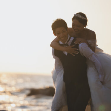 Smiling groom giving the bride a piggyback ride by the sparkling sea, a playful pose for seaside wedding photography on the Chita Peninsula, Aichi