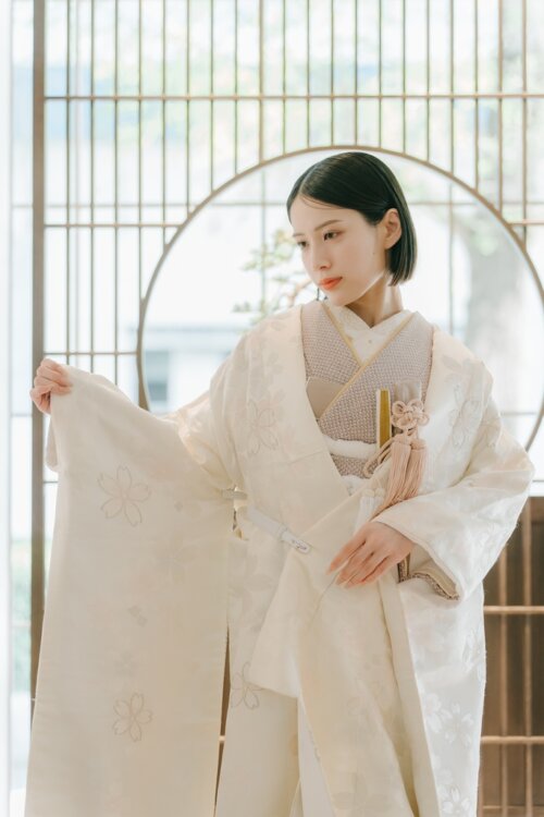 Bride in a shiromuku spreading her sleeve to show the cherry blossom embroidery in a Japanese room with a round window and lattice doors, a kimono wedding photo in Japan (Shiromuku AS102).