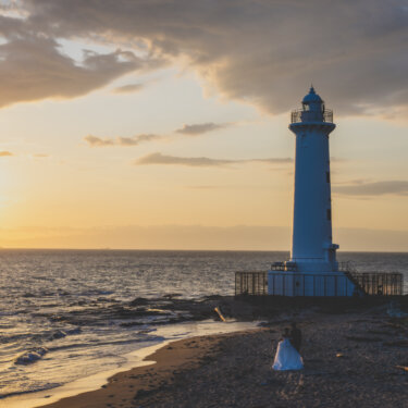 Noma Lighthouse standing against a dramatic sunset sky with the couple small on the beach below, a scenic wedding photoshoot spot in Aichi, Japan