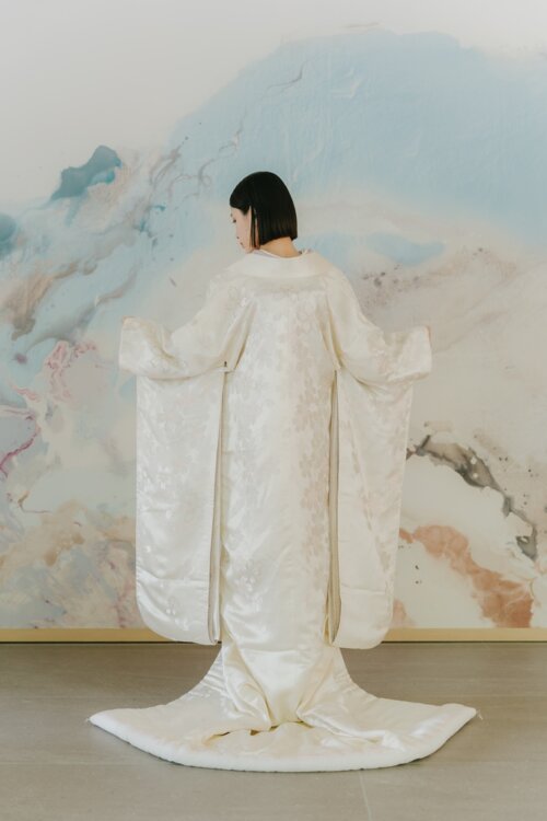 Back view of a bride in a shiromuku spreading both sleeves in front of a pale blue mural, showing the woven pattern from shoulders to train in this Japan wedding photography shot (AS102).
