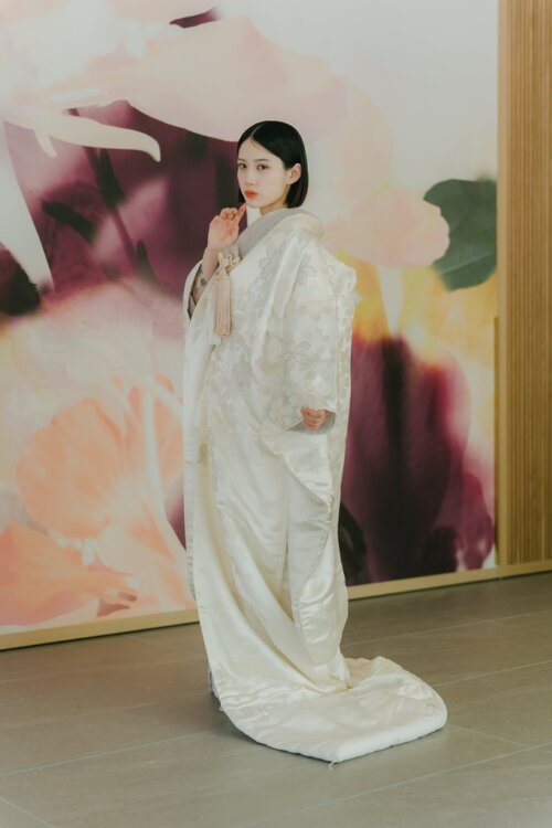 Side view of a bride in a shiromuku holding the tassel of her hakoseko against a floral wall, a full-length studio photo for a pre-wedding photoshoot in Japan (Shiromuku AS102).