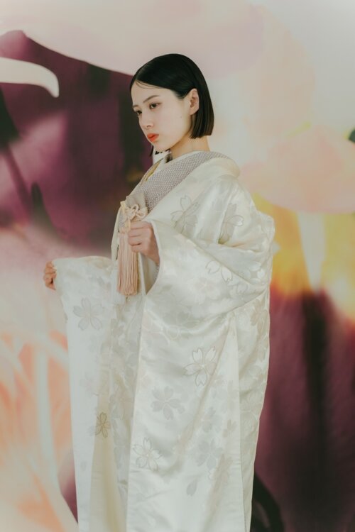 Bride lifting her shiromuku sleeve to reveal the cherry blossom pattern in front of a large floral art wall with dusty pink tassels, kimono wedding photos in Japan (AS102).