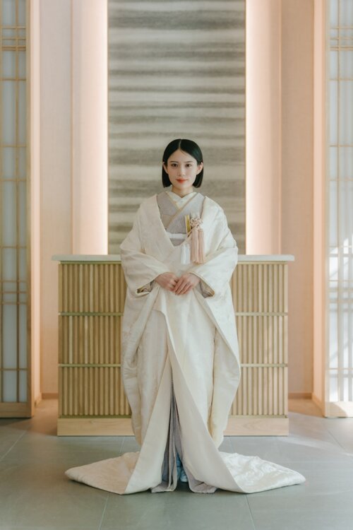 Bride in a shiromuku standing front-facing with hands folded in a modern Japanese-style studio, the full length of the kimono shown for a pre-wedding photoshoot in Japan (Shiromuku AS104).