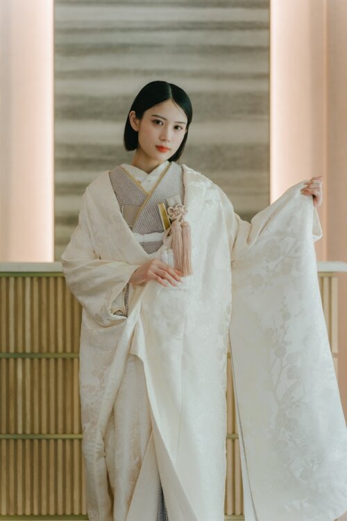 Bride opening one shiromuku sleeve to show the woven pattern beside a lattice and wooden counter in a modern Japanese studio, kimono wedding photos in Japan (AS104).