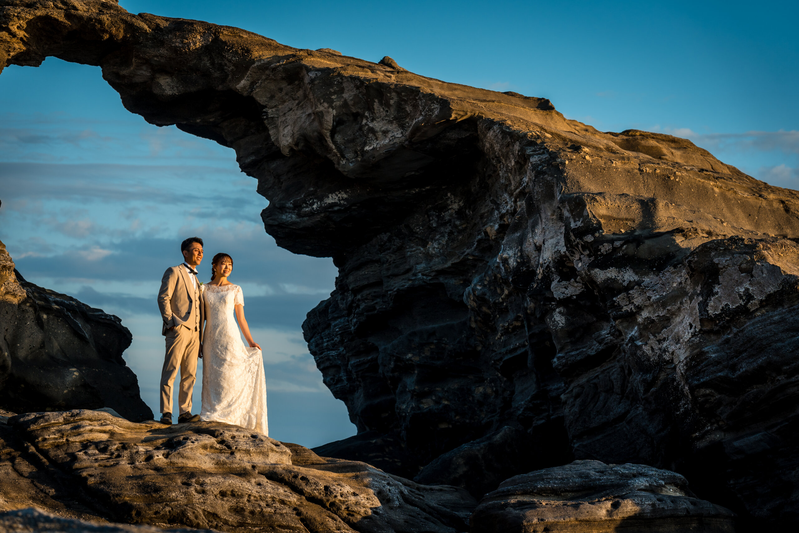 Bride and groom standing under the natural rock arch of Umanose Domon on Jogashima, with the sea and blue sky visible through the arch