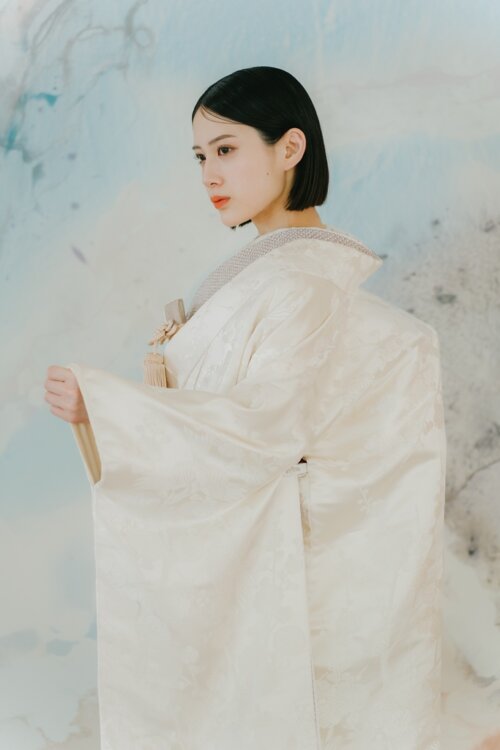 Bride in a shiromuku spreading her sleeve and turning her profile toward the camera in front of a pale blue mural, a popular kimono for pre-wedding photoshoots in Japan (AS104).