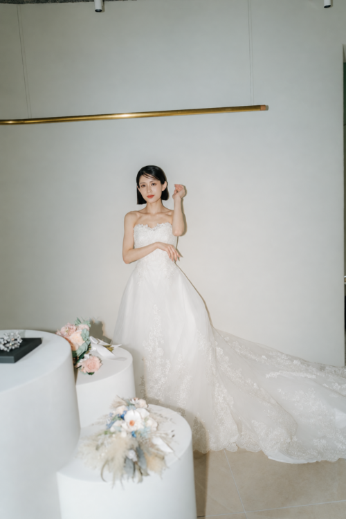 Bride in a bustier wedding dress touching her hair beside a white column with a dried flower bouquet during a studio wedding photoshoot in Japan