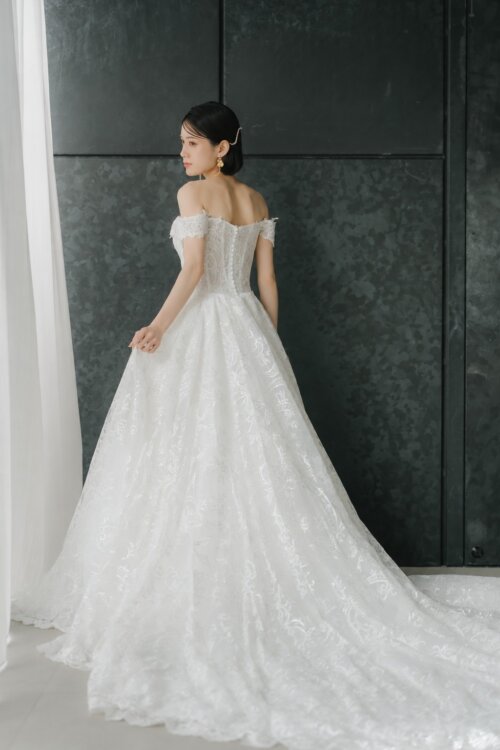 Bride in an off-shoulder all-lace wedding dress looking back over her shoulder with the long train spread on the floor, a back view for a pre-wedding photoshoot in Japan
