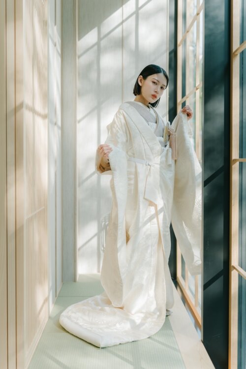 Bride in a shiromuku lifting her sleeve and spreading the hem in a corridor of latticed windows with light and shadow, a natural-light studio photo for a pre-wedding photoshoot in Japan (Shiromuku AS105).