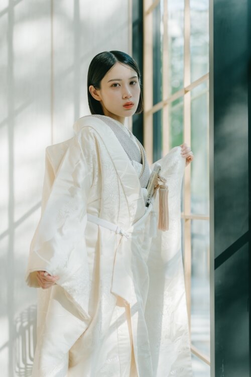 Bride in a shiromuku spreading her sleeves and looking at the camera as light pours through latticed windows, pink tassels accenting this kimono wedding photo in Japan (AS105).