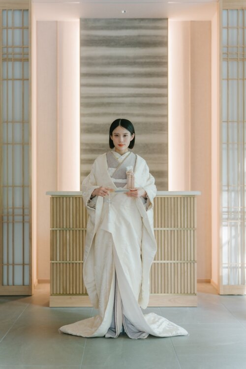 Bride in a shiromuku standing at the center of a modern Japanese studio framed by shoji and lattice, holding a fan in a symmetrical composition, Japan wedding photography (AS105).