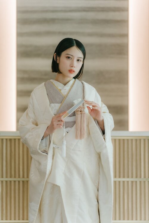 Bride in a shiromuku facing forward with a fan and pink tassels in a modern Japanese space with lattice and soft lighting, an upper-body pre-wedding photoshoot in Japan (Shiromuku AS105).