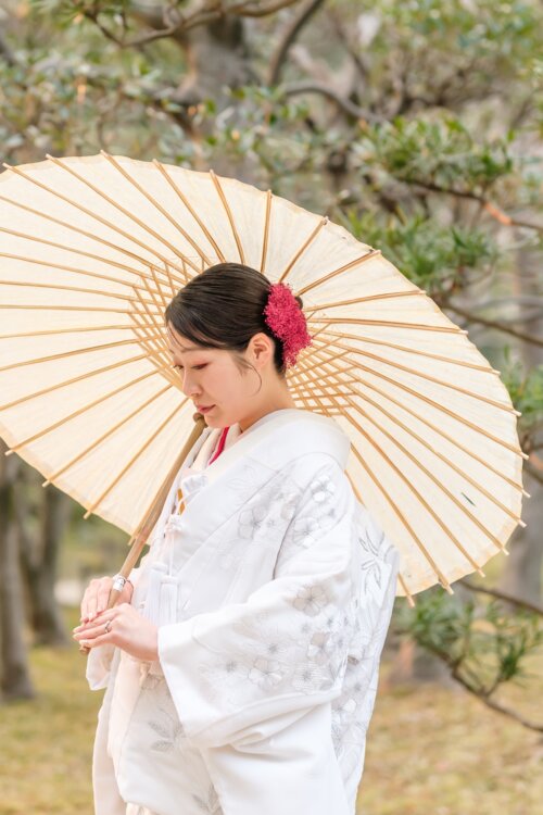 Bride in shiromuku with a red hair ornament holding a wagasa umbrella under the trees of a Japanese garden, Japan wedding photography