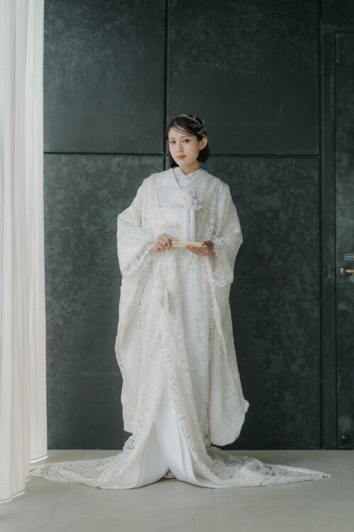 Bride in a lace-patterned shiromuku holding a folding fan before a dark stone wall, a full-length studio photo for a pre-wedding photoshoot in Japan (Shiromuku DE101).